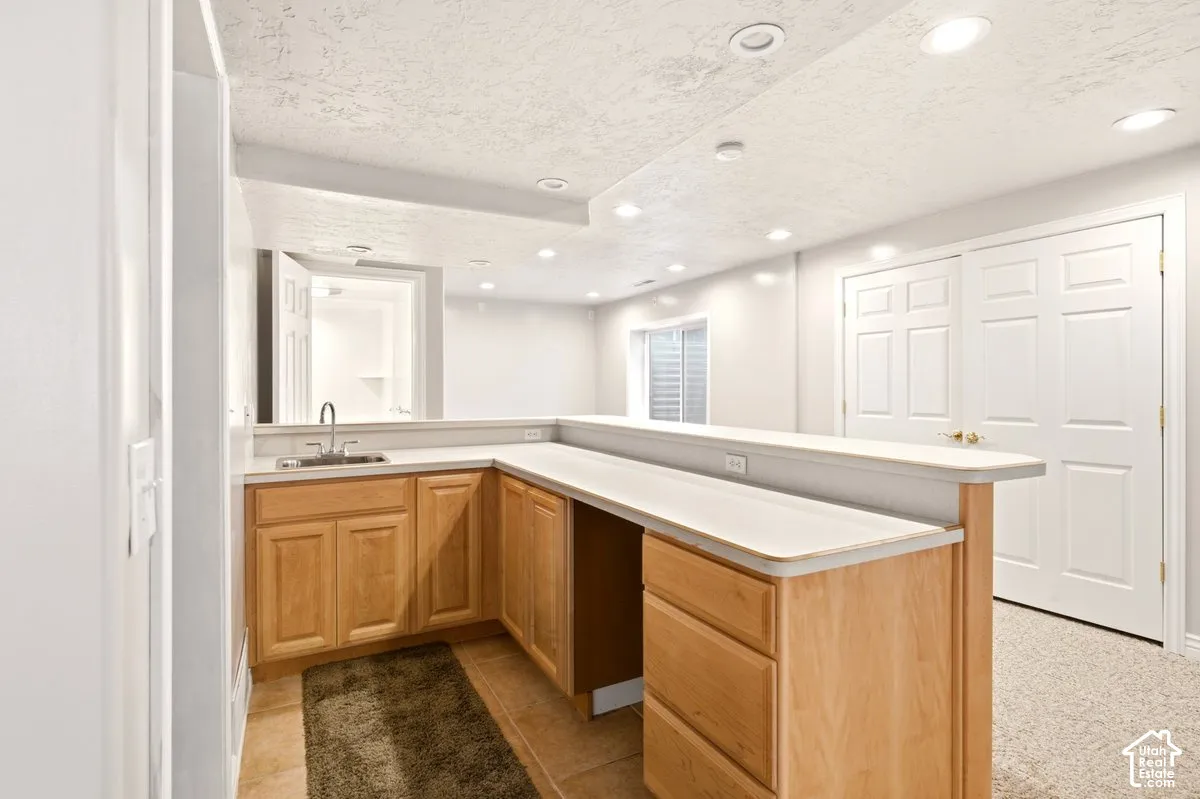 L-shaped kitchenette featuring light-wood cabinetry, a stainless steel sink, a neutral-toned countertop with integrated breakfast bar, and recessed ceiling lighting