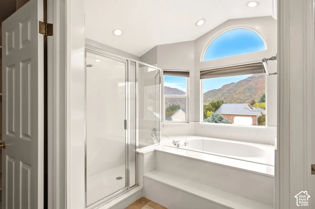 Glass-enclosed shower, soaking tub with tile surround, vaulted ceiling with recessed lighting, and arched window with mountain backdrop
