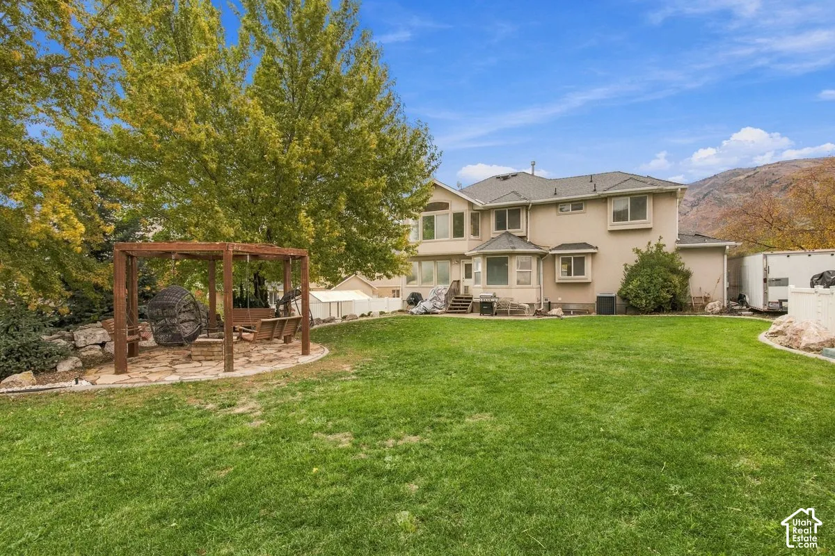 Expansive backyard featuring a wood-frame pergola, stone-paved patio, manicured lawn, mature trees, and mountain backdrop