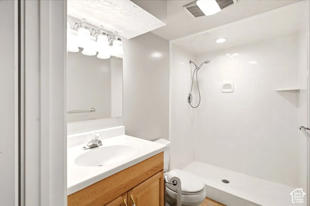 Bathroom featuring a wood-finish vanity with a white integrated countertop and basin, five-bulb vanity lighting fixture, wall-mounted mirror, and walk-in shower with a handheld showerhead and built-in shelving