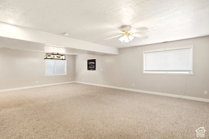 Spacious room featuring neutral carpeting, a ceiling fan with integrated lighting, dual windows with blinds, and a decorative hanging light fixture