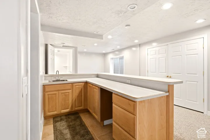 L-shaped kitchenette featuring light-wood cabinetry, a stainless steel sink, a neutral-toned countertop with integrated breakfast bar, and recessed ceiling lighting