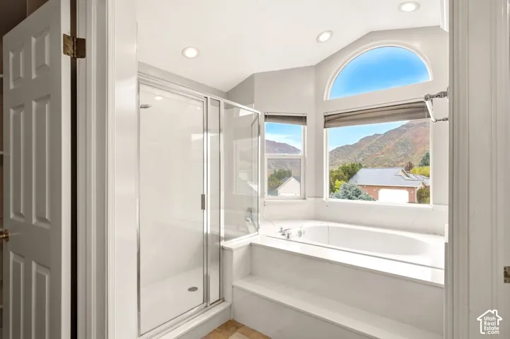 Glass-enclosed shower, soaking tub with tile surround, vaulted ceiling with recessed lighting, and arched window with mountain backdrop
