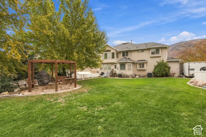 Expansive backyard featuring a wood-frame pergola, stone-paved patio, manicured lawn, mature trees, and mountain backdrop