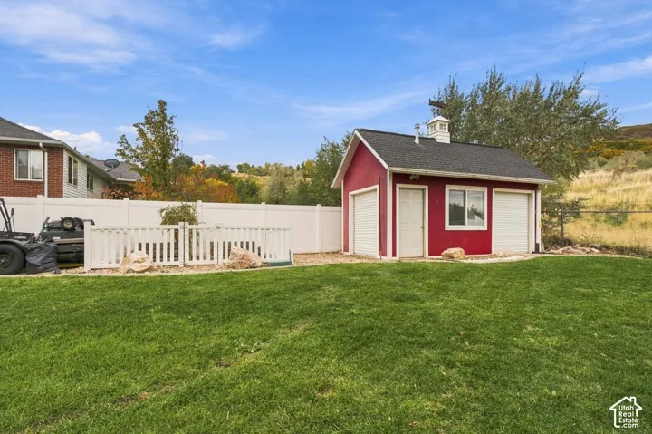 Detached outbuilding featuring a red exterior, white trim, dual roll-up doors, a single entry door, and a cupola, situated on a lot with a white vinyl fence and manicured lawn