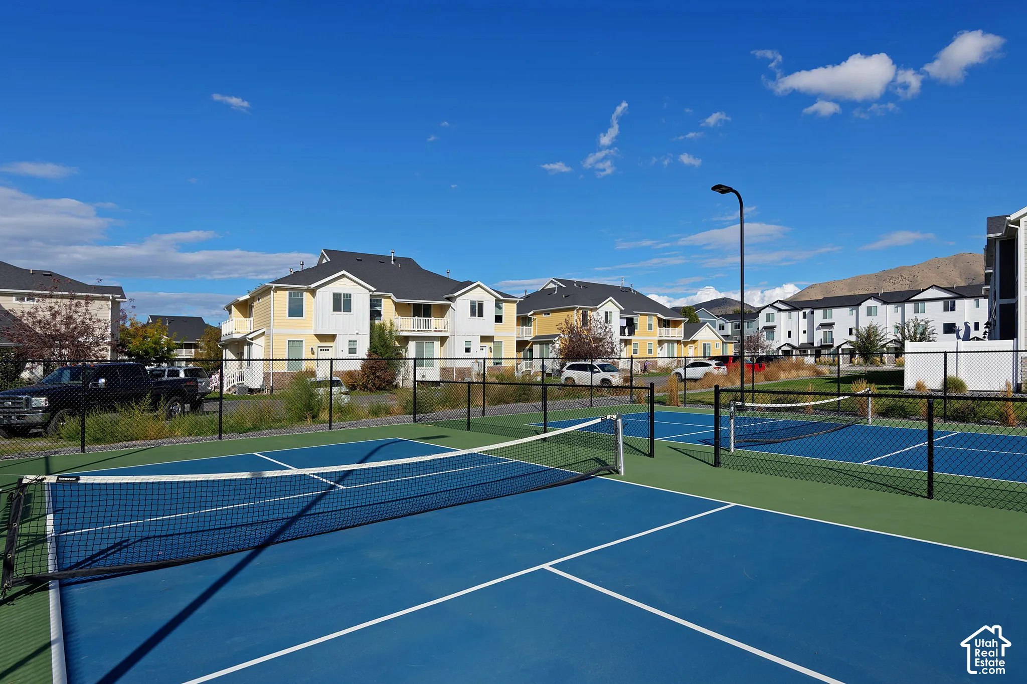 Hard-surfaced pickleball courts enclosed by black metal fencing, featuring integrated nets and pole-mounted lighting