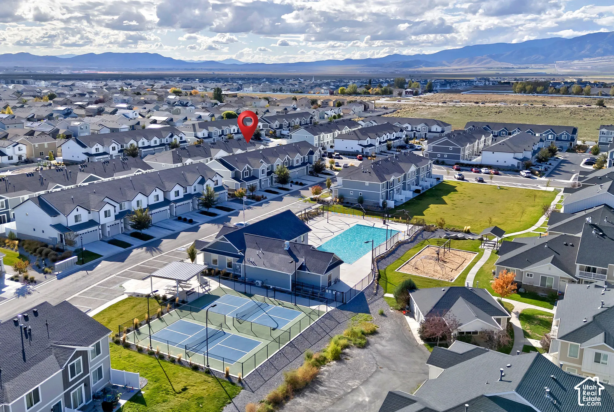 Aerial perspective of a community complex featuring an outdoor swimming pool, fenced tennis courts, and a playground area, set against a mountain backdrop