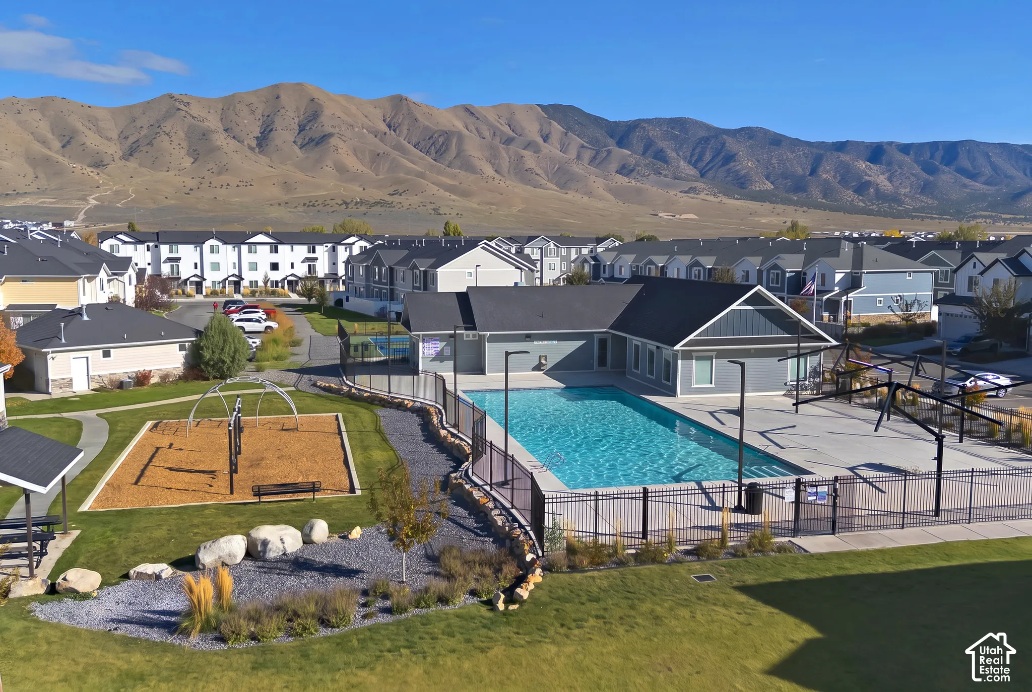 Community recreation area featuring a fenced swimming pool, paved pool deck, playground with wood-chip surfacing, and mountain backdrop