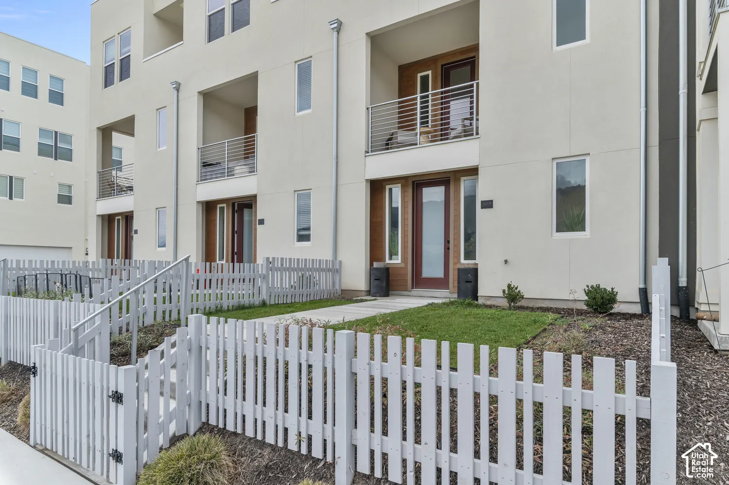 Multi-level exterior featuring light-toned stucco siding, private balconies with horizontal metal railings, wood-paneled entry accents, and a fenced front yard with a concrete walkway