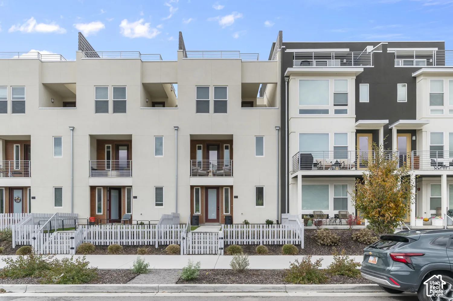 Modern multi-level exterior featuring clean-lined stucco and dark paneling, private balconies with cable railings, and individual white picket-fenced front entries