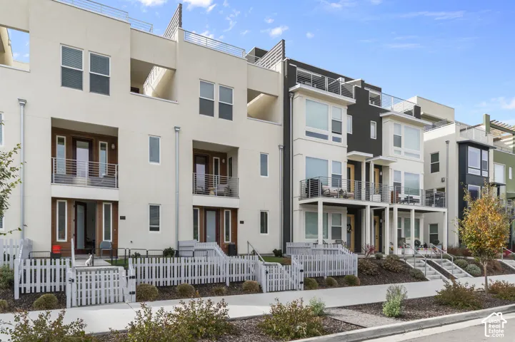 Modern multi-level exterior featuring light and dark contrasting stucco facades, metal terrace railings, recessed private balconies, and white picket-fenced front entryways