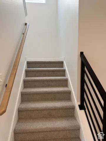 Staircase featuring neutral carpeted treads, a light wood-finish wall-mounted handrail, and a matte black metal horizontal-bar railing