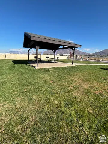 Covered pavilion with a shingle roof over a concrete slab, set within a grass-covered lot featuring mountain backdrops
