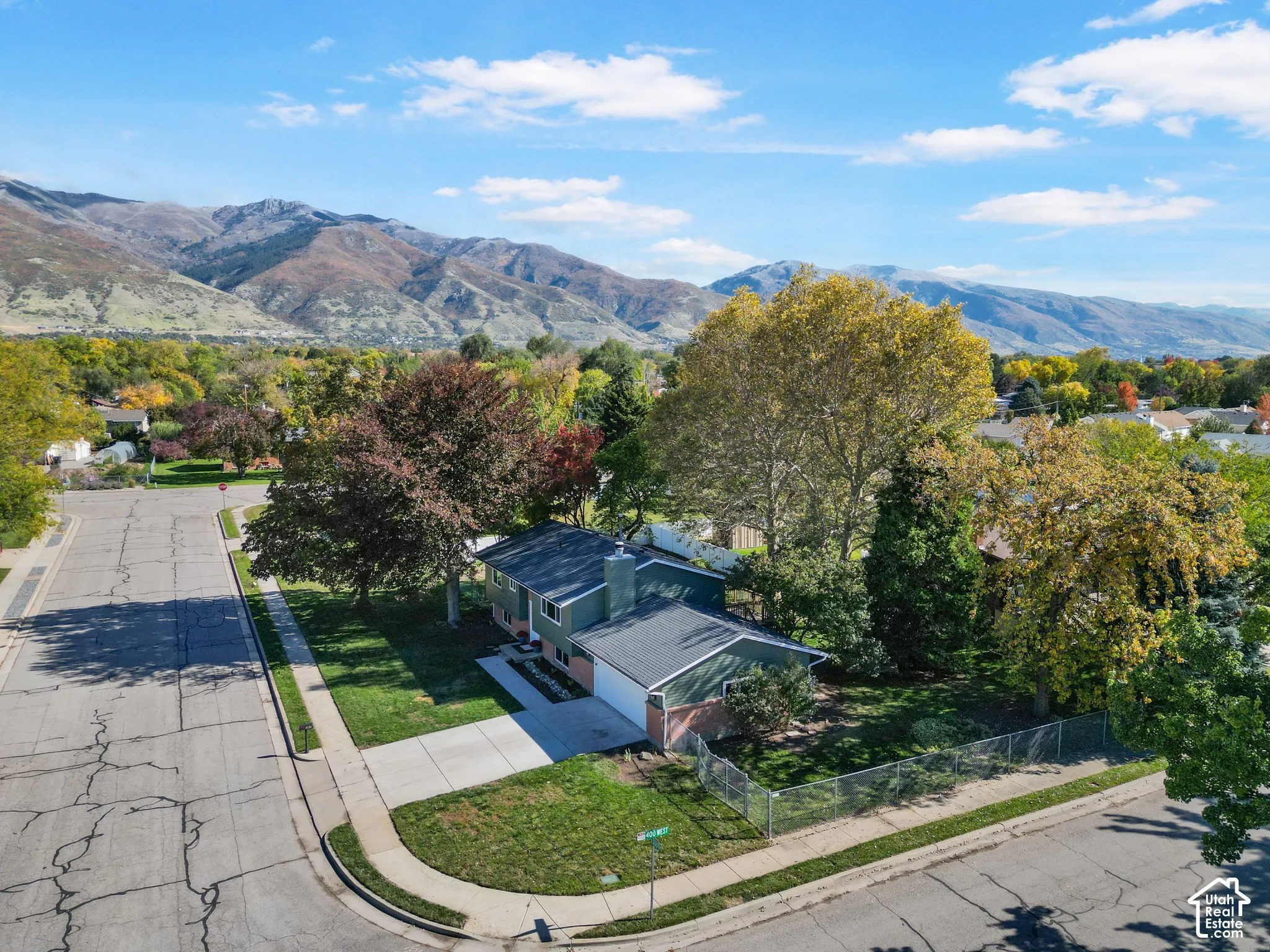 Corner lot residence featuring a two-car garage, concrete driveway, chain-link fencing, and mature trees against a mountain backdrop
