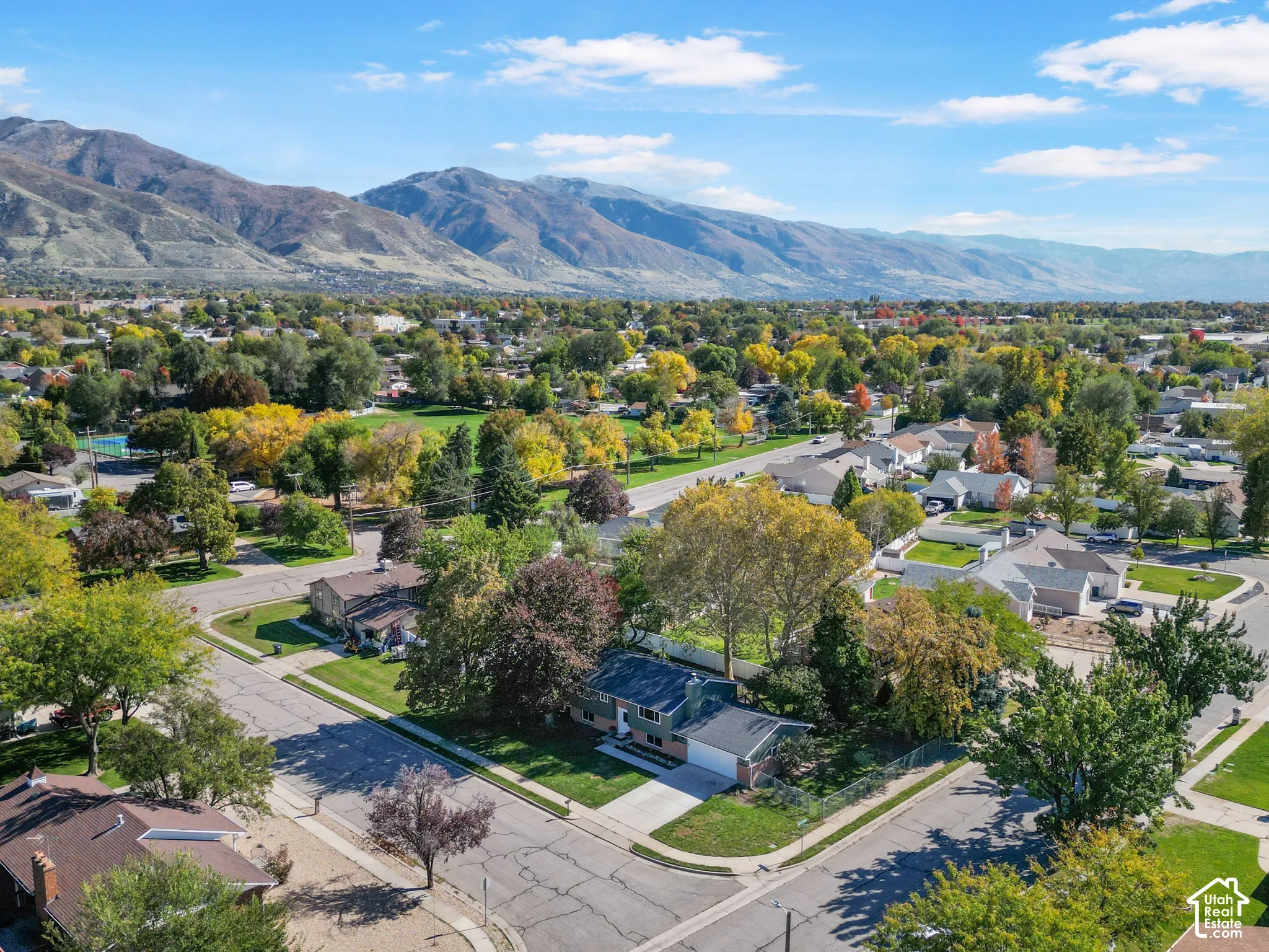 Aerial perspective featuring a residential property with a two-car garage, concrete driveway, mature trees, and prominent mountain backdrops