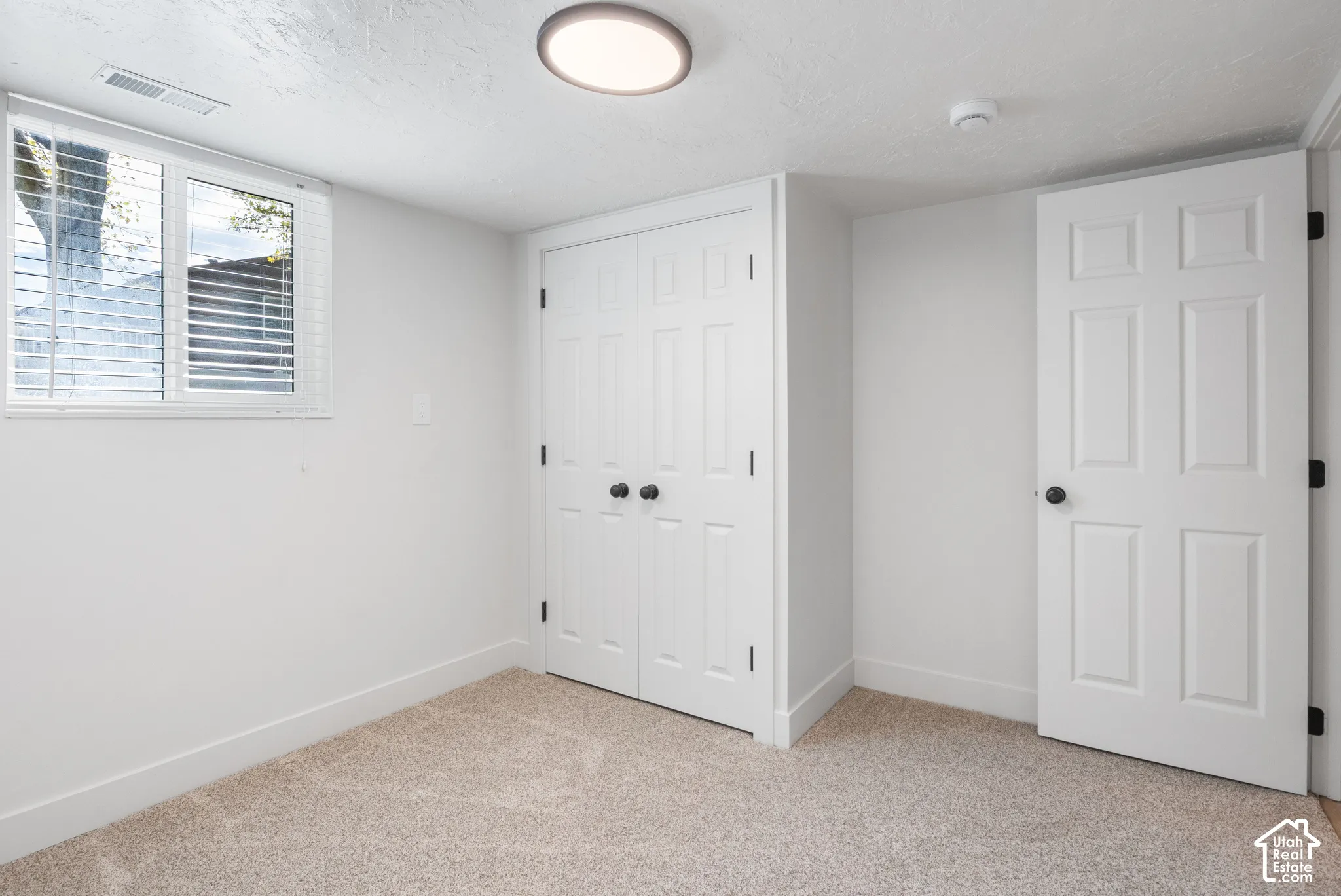 Room featuring neutral carpeting, double-door closet with round hardware, six-panel interior door, and natural light from a window with horizontal blinds