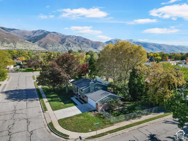 Corner lot residence featuring a two-car garage, concrete driveway, chain-link fencing, and mature trees against a mountain backdrop