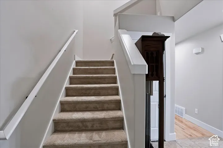 Staircase featuring neutral carpeted treads, white handrails, and wood-finish flooring in the hallway