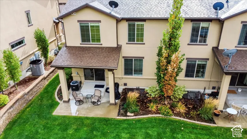 Stucco exterior featuring a covered rear patio, concrete hardscaping, landscaped garden beds with mature plantings, and a manicured lawn