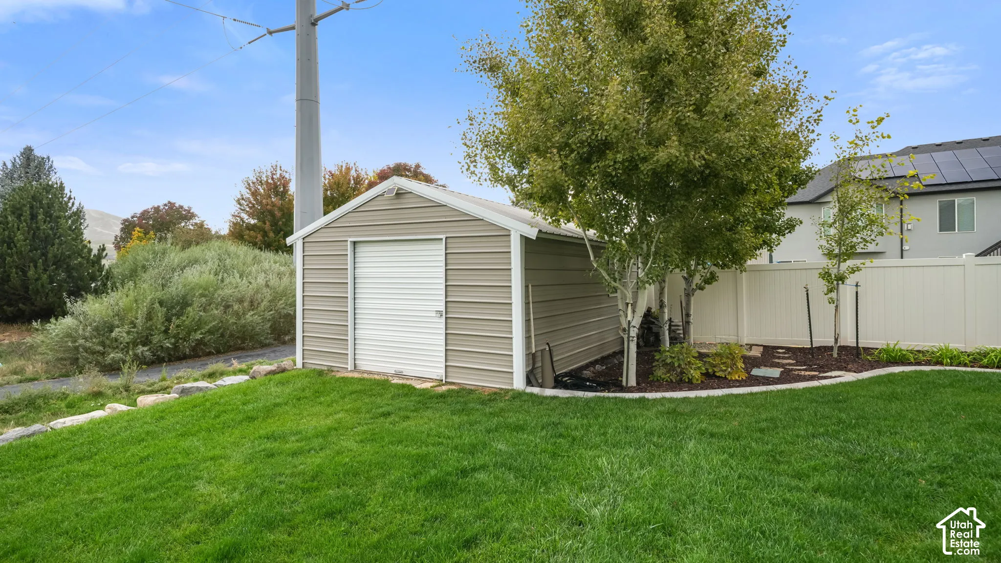 Detached metal storage shed featuring a roll-up door, set on a manicured lawn with mature trees and white vinyl privacy fencing