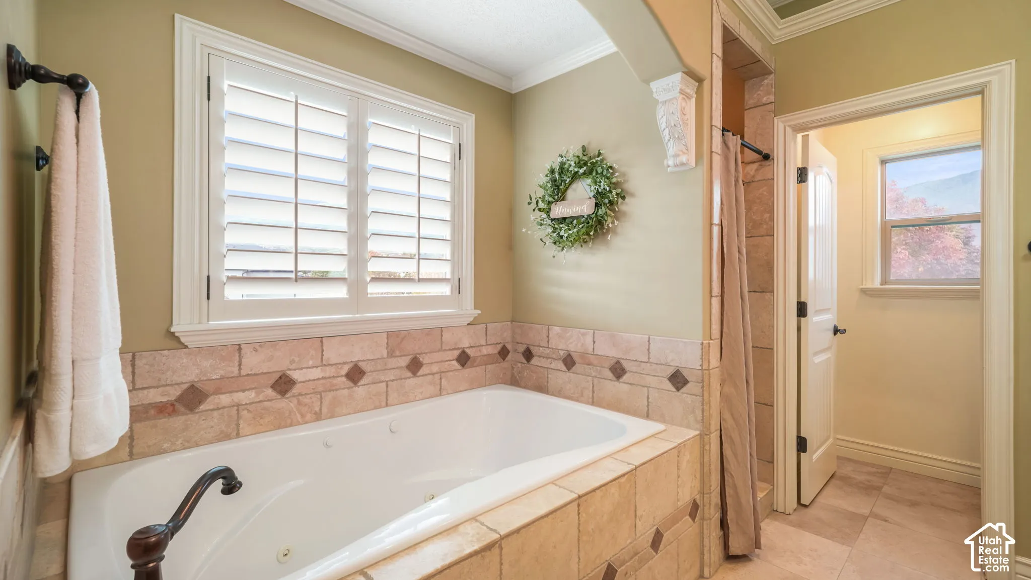 Jetted soaking tub featuring tile surround with diamond-patterned inlay, white plantation shutters, decorative corbel, and oil-rubbed bronze fixture