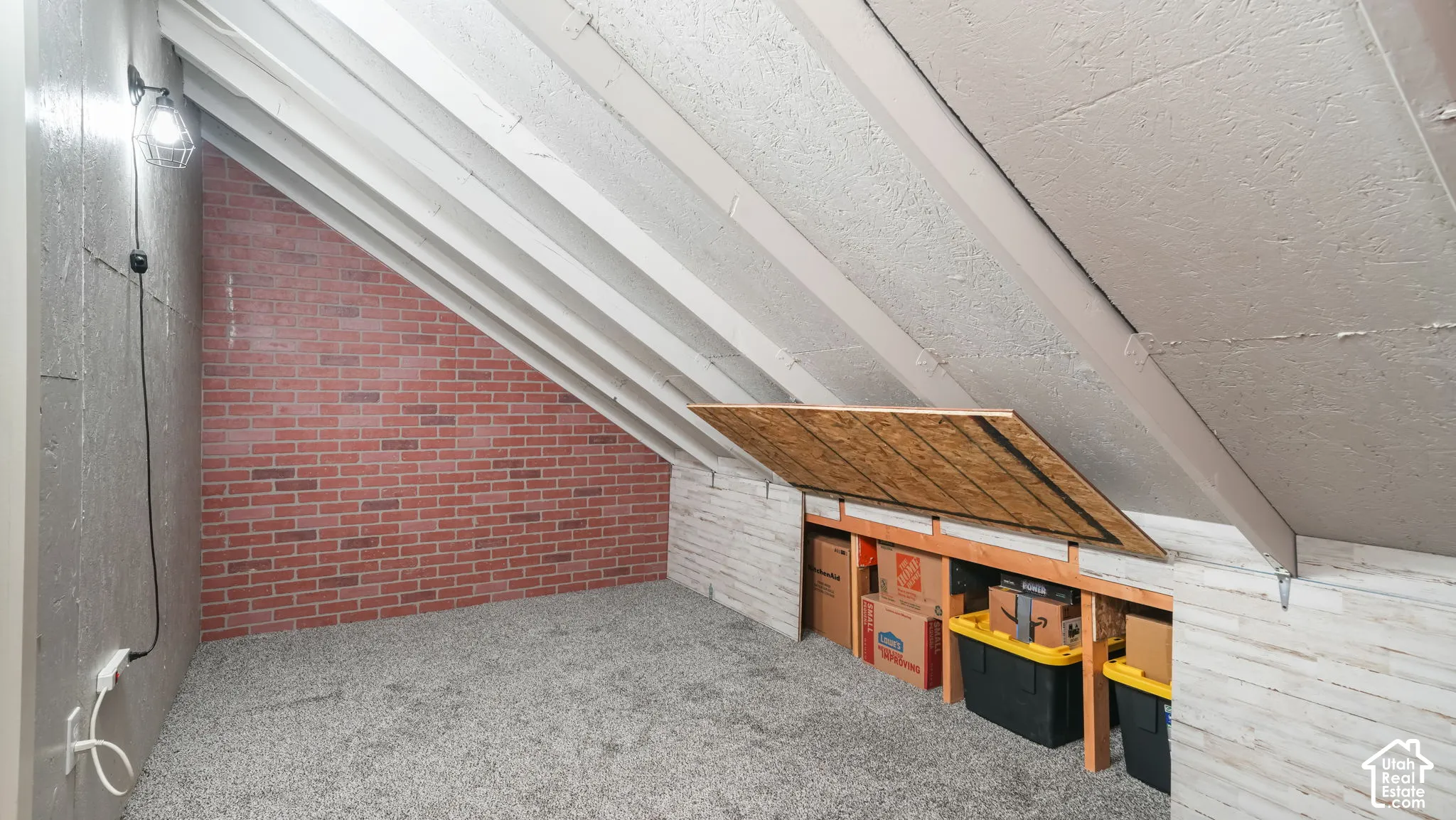 Attic space featuring a red brick accent wall, neutral carpeting, exposed rafters, and integrated knee-wall storage access