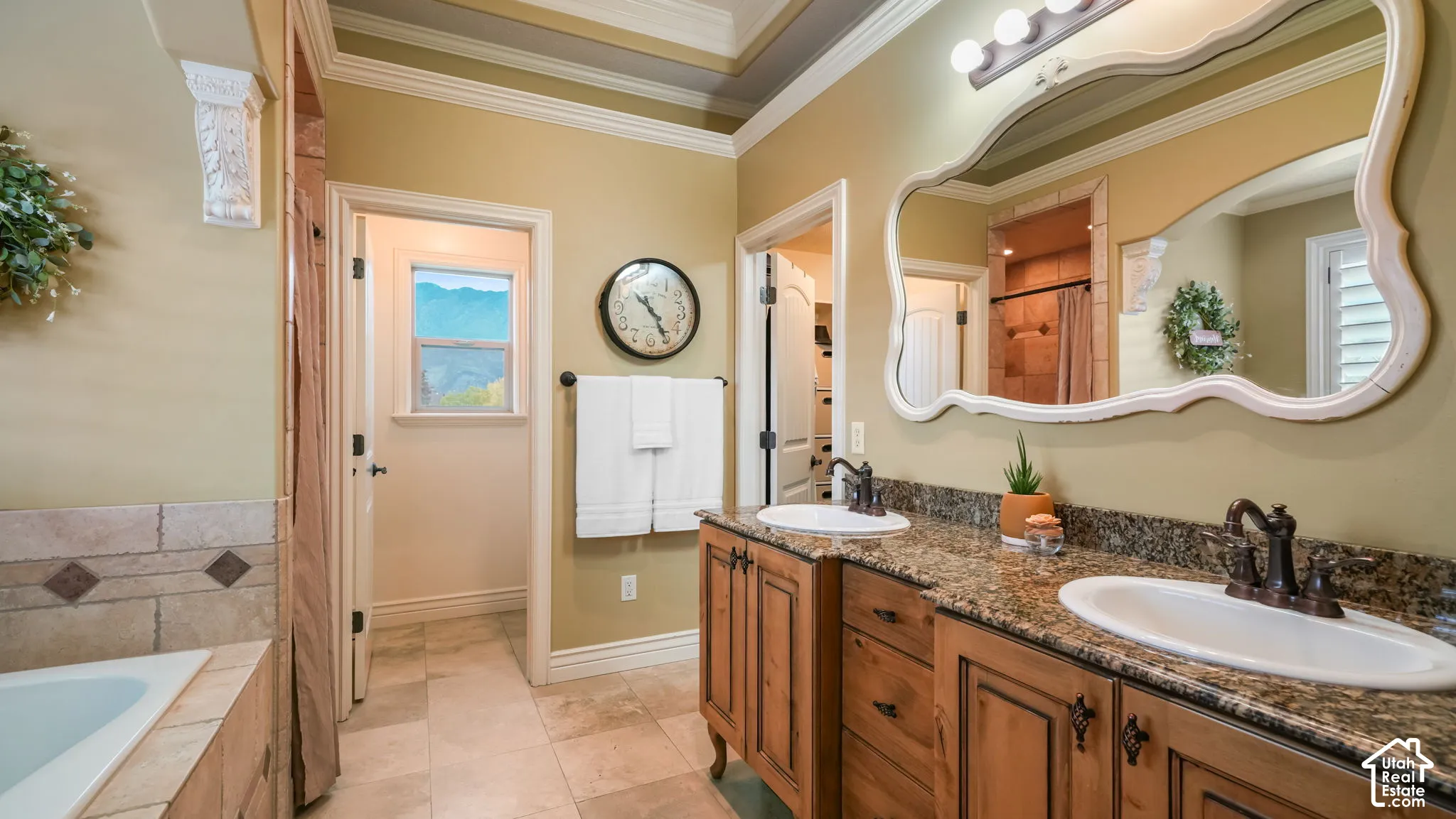 Dual-vanity cabinetry with stone countertops, undermount sinks, bronze-finish fixtures, and a decorative wall mirror, complemented by tile flooring and a tray ceiling