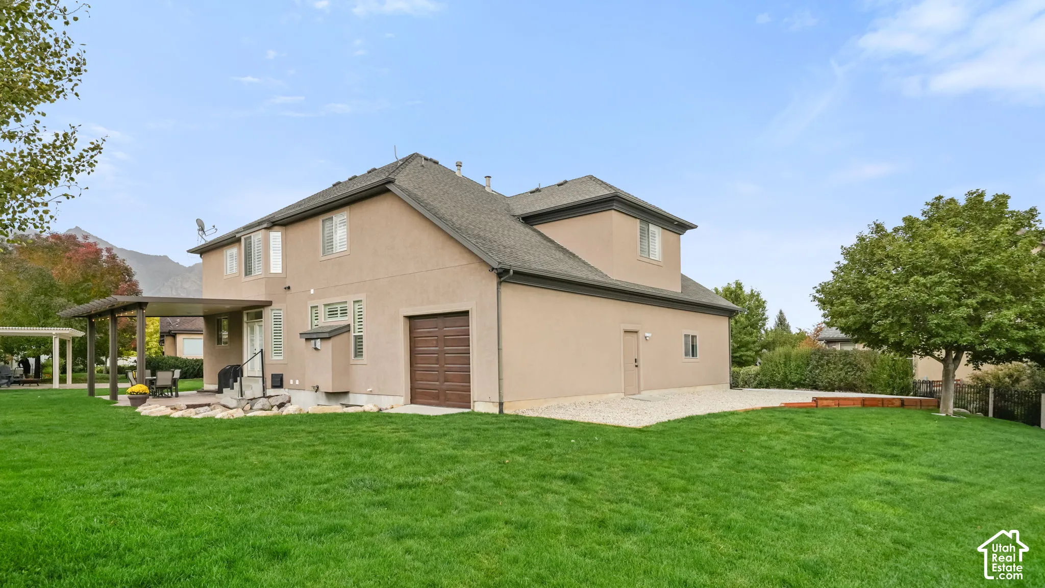Exterior featuring a stucco facade, attached garage with wood-finish door, covered patio, and expansive lawn with mature landscaping