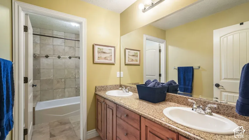 Bathroom featuring a dual-sink vanity with stone-surfaced countertops, wood cabinetry, a large wall-mounted mirror, and a tub-shower combination with decorative tile surround