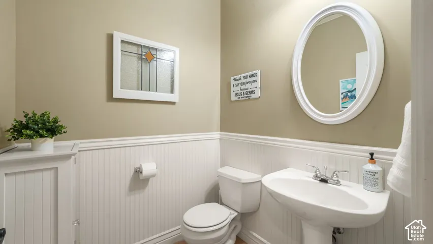 Bathroom featuring white beadboard wainscoting, a wall-mounted oval mirror with a white frame, a porcelain pedestal sink with polished chrome hardware, and an inset decorative window