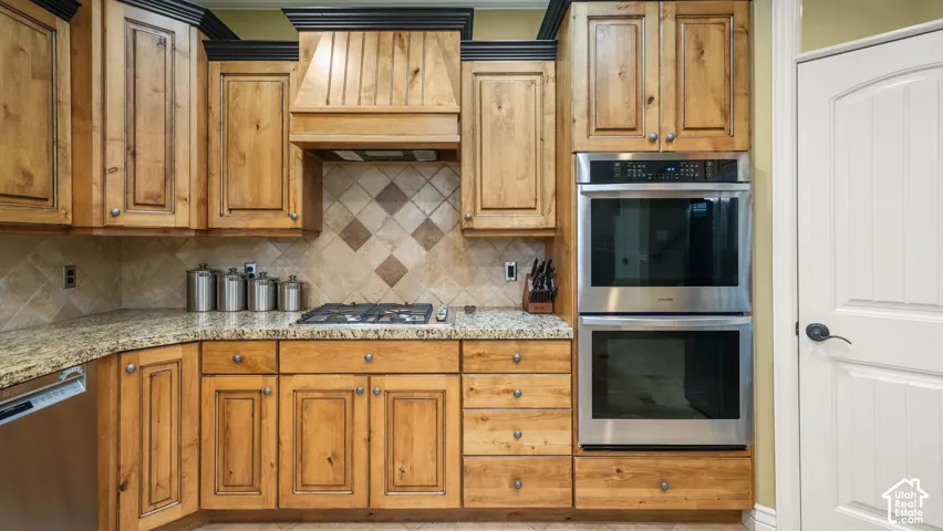 Custom cabinetry with matching range hood, stone countertops, diamond-pattern tile backsplash, stainless steel built-in double ovens, and integrated gas cooktop