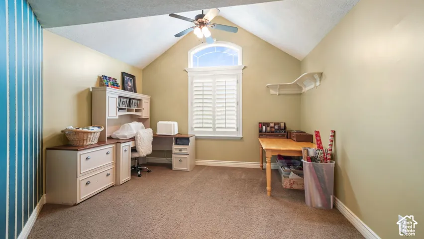 Vaulted ceiling room featuring a central arched window with plantation shutters, a ceiling fan with integrated light fixture, carpeted flooring, and a wall-mounted floating shelf
