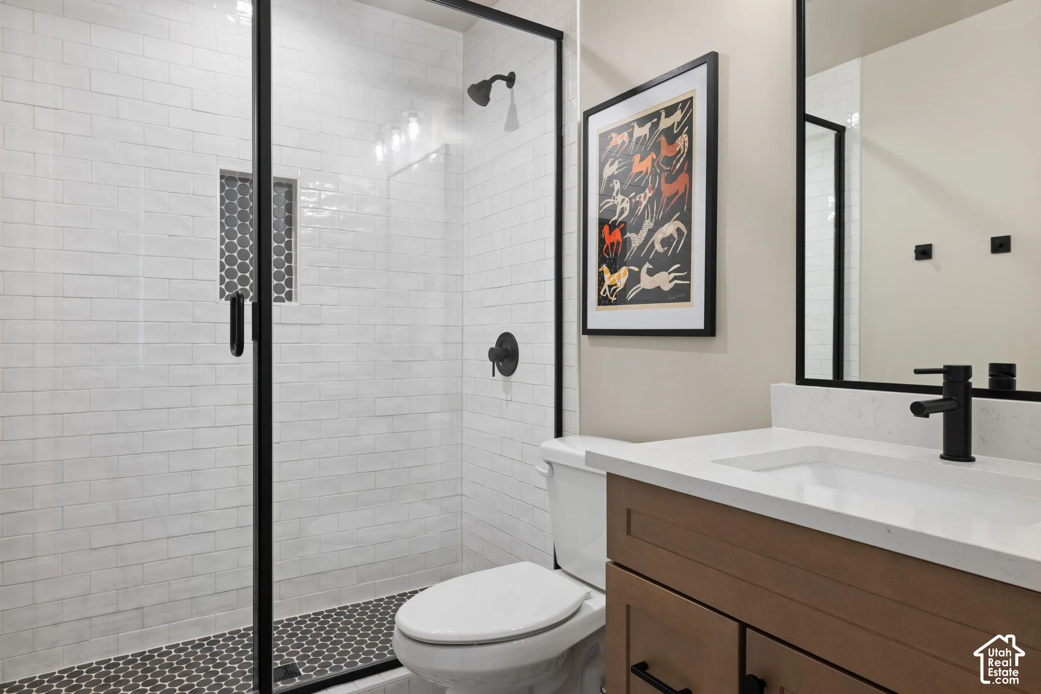 Bathroom featuring a vanity with quartz countertop, under-mount sink, and matte black fixtures, paired with a glass-enclosed shower, white subway tile surround, and mosaic tile flooring