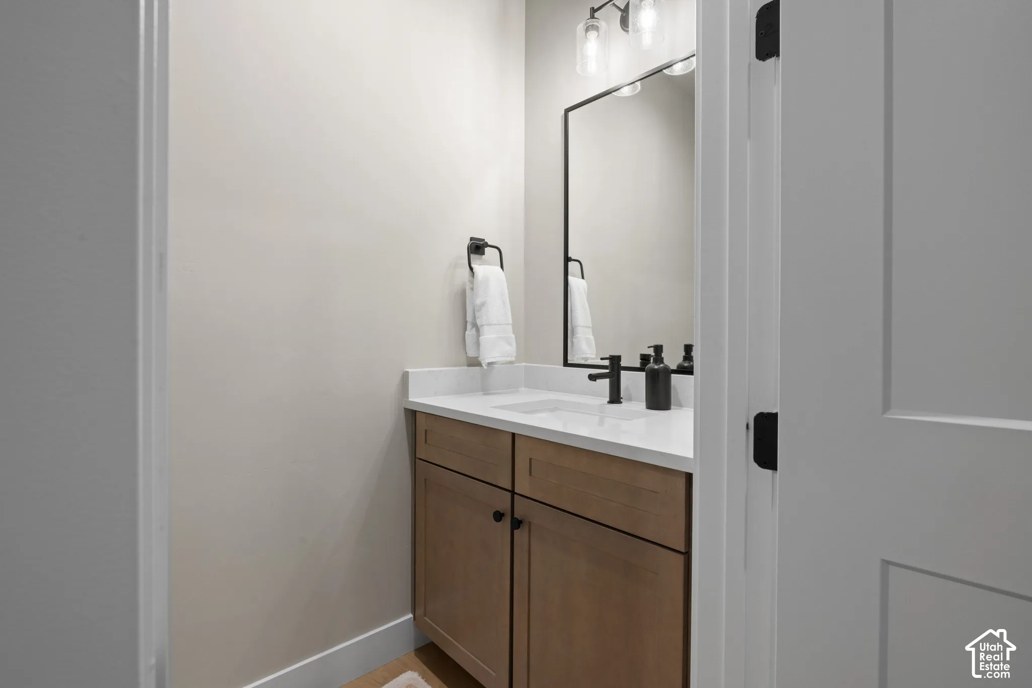 Cabinetry with a light wood finish, white countertop with an integrated rectangular sink, matte black faucet, rectangular wall mirror with a black frame, and three-light glass fixture