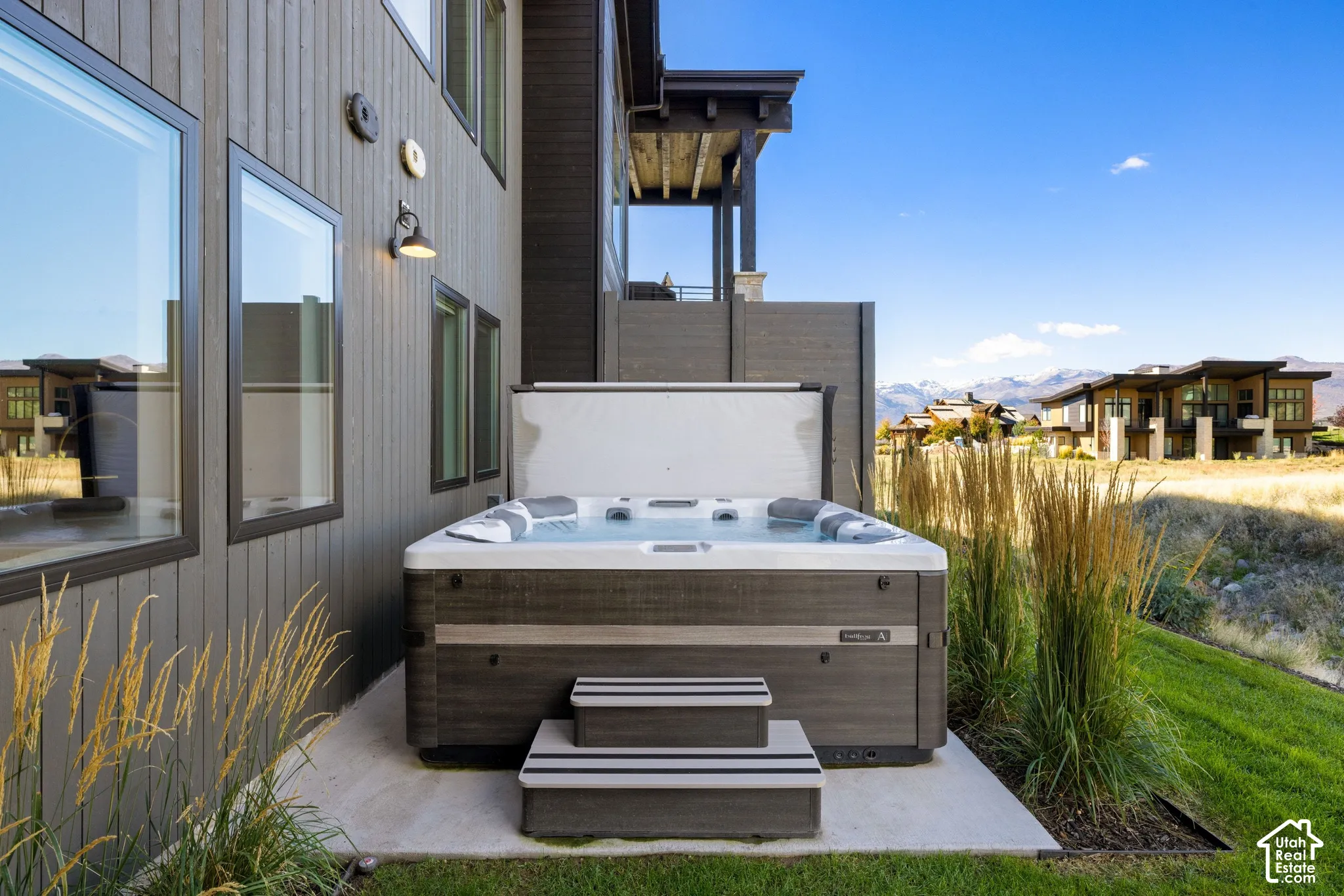 Hot tub with integrated steps situated on a concrete patio, vertical wood-finish siding, exterior wall-mounted lighting, and mountain scenery