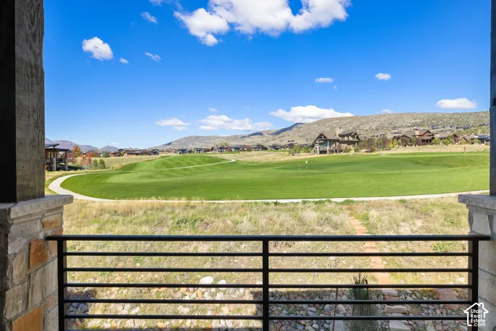 Covered stone-accented patio featuring a metal horizontal-slat railing, overlooking manicured golf course grounds and distant mountain terrain