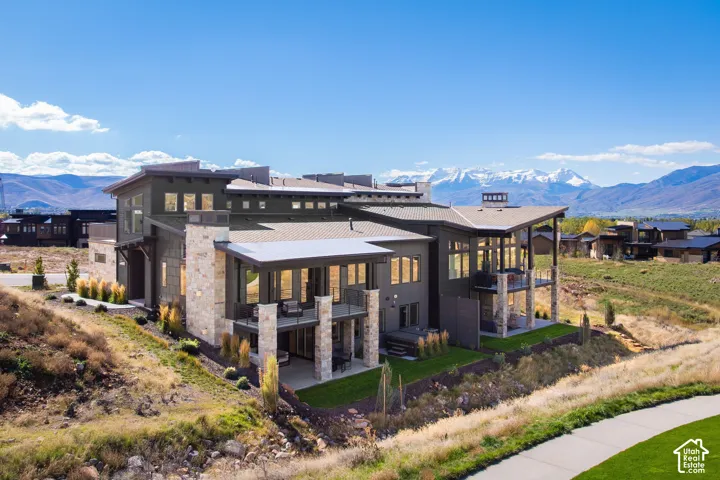 Modern multi-level exterior featuring stacked stone columns, expansive upper-level balconies, ground-level covered patios, and multi-pitch rooflines set against a mountain backdrop