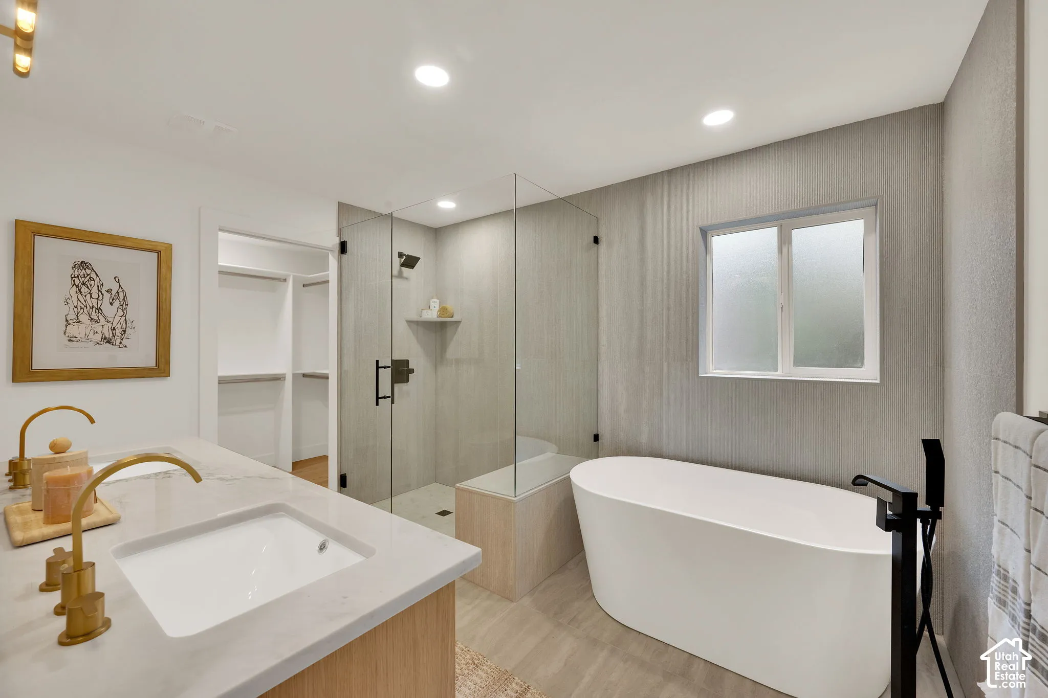 Primary bathroom featuring a free-standing soaking tub with matte black floor-mounted faucet, walk-in glass-enclosed shower featuring stone-finish wall tile, dual-sink vanity with light-tone stone countertop and gold-finish hardware, and recessed ceiling
