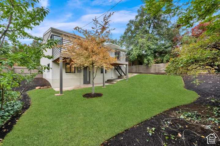 Exterior featuring a elevated metal balcony with horizontal railing, concrete patio, staircase with metal railings, landscaped lawn, and mature trees