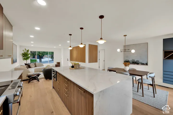 Kitchen featuring a waterfall-edge island with light-toned surfacing, wood-finish cabinetry, recessed lighting, decorative pendant fixtures, and wood flooring