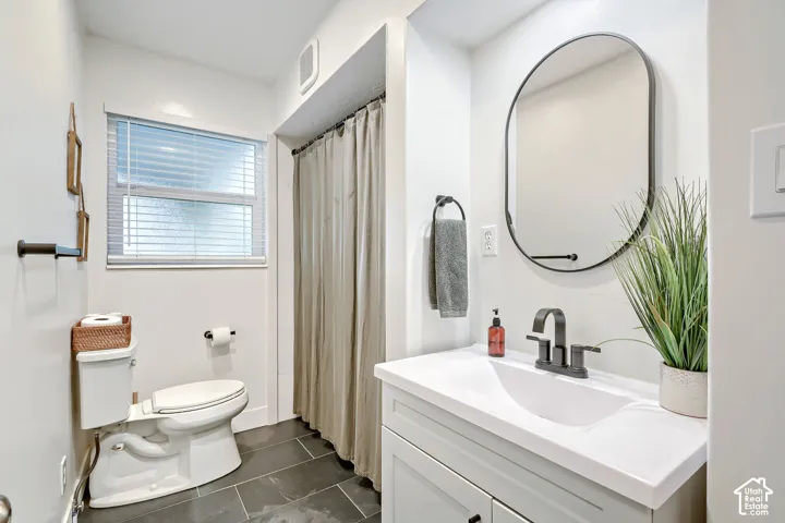 Bathroom featuring a vanity with an integrated basin, matte black faucet, oval-shaped mirror, dark tile flooring, and a window with blinds