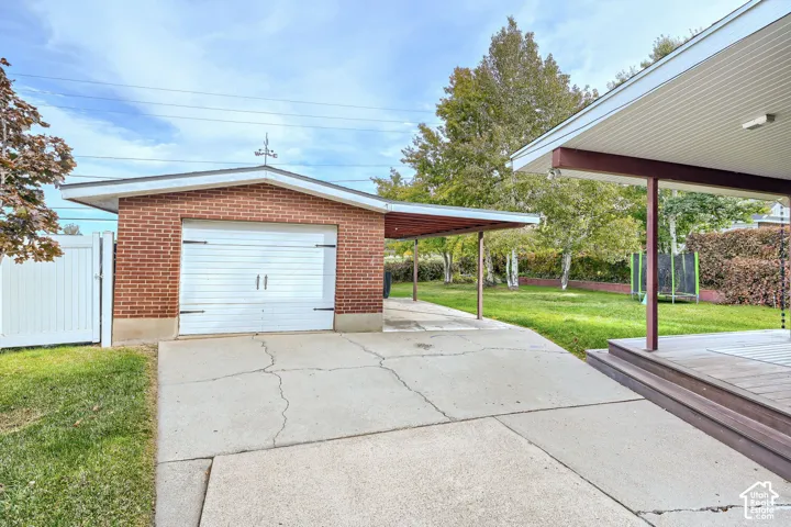 Brick detached garage featuring a single-car overhead door, covered carport extension, concrete driveway, and white vinyl perimeter fencing