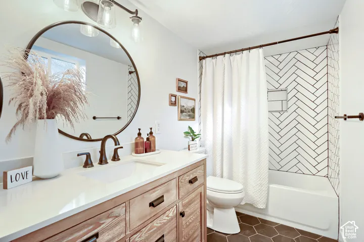 Wood-finish cabinetry with a solid surface countertop, round metal-frame mirror, herringbone-patterned tile shower surround, and dark hexagonal floor tile