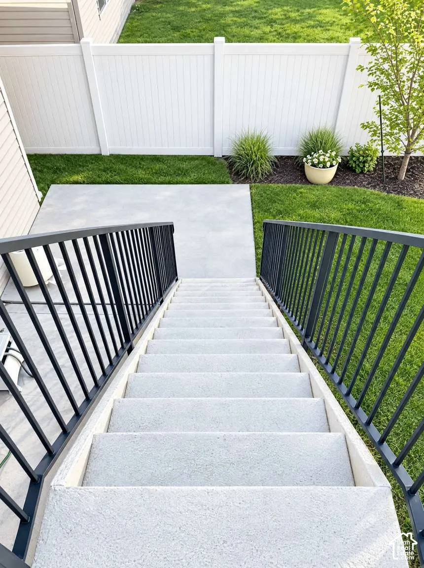 Concrete staircase featuring black metal railings leading to a concrete patio, enclosed by a white vinyl privacy fence and landscaped lawn