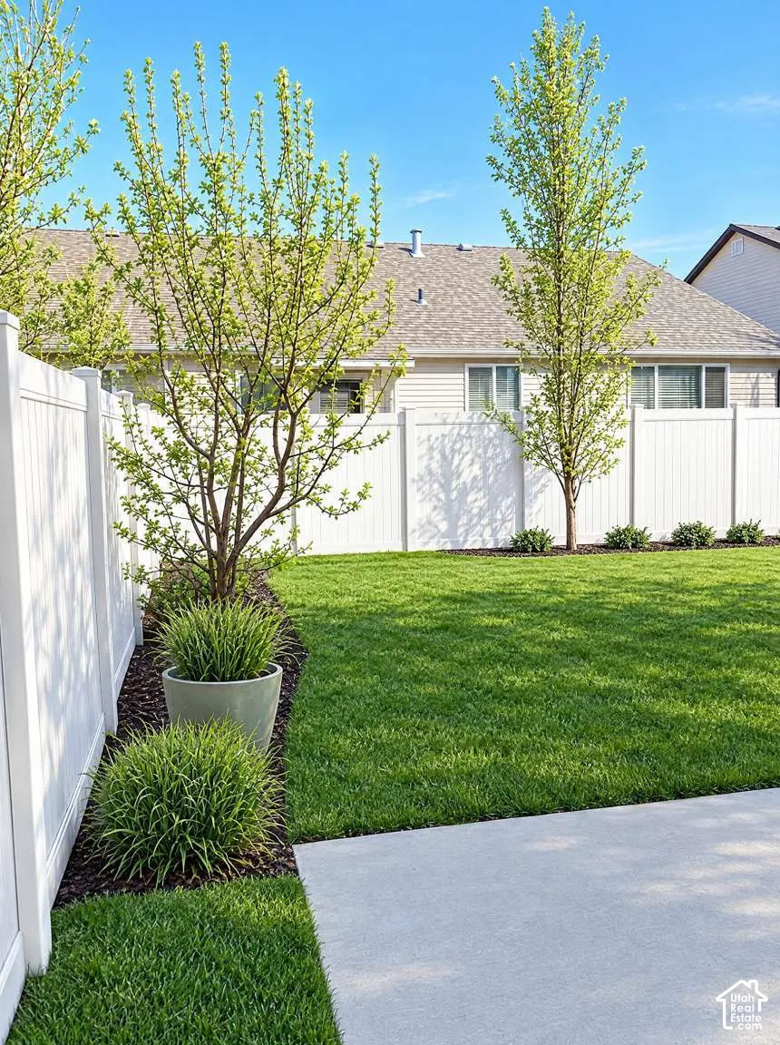Fenced backyard featuring a manicured lawn, concrete patio, established trees, and dark mulch garden beds