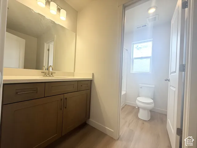 Bathroom featuring a wood-finish vanity with a light-toned countertop, large frameless mirror, three-light fixture, and wood-finish flooring
