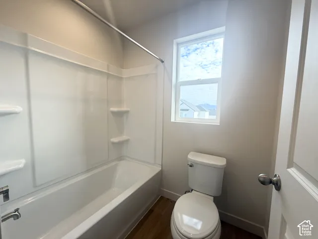 Bathroom featuring a white tub-shower combination with integrated shelving, a dual-flush toilet, wood-finish flooring, and a single-hung window
