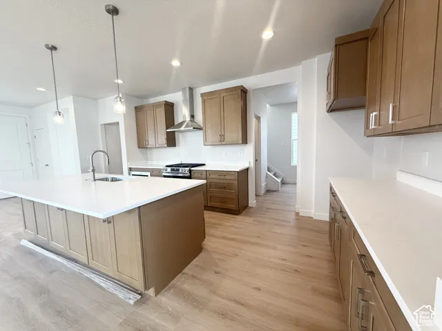Kitchen featuring wood-finish cabinetry, white countertops, a center island with an undermount sink, stainless steel range hood, and wood-finish flooring