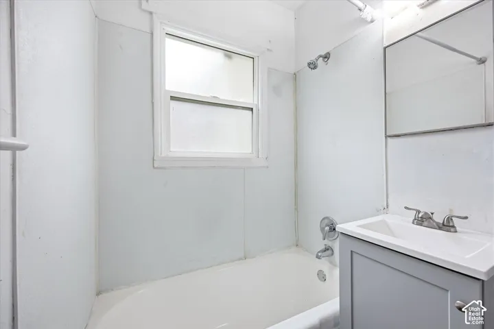 Bathroom featuring a bathtub with a wall-mounted shower head, a single-basin vanity with a grey cabinet, a rectangular mirror, and a window with white trim