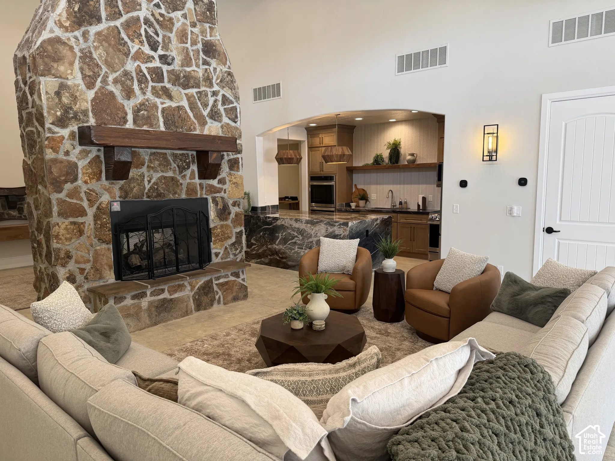 Floor-to-ceiling natural stone fireplace with a thick wood mantel, vaulted ceilings, and a kitchen featuring a large slab-stone waterfall island and built-in cabinetry